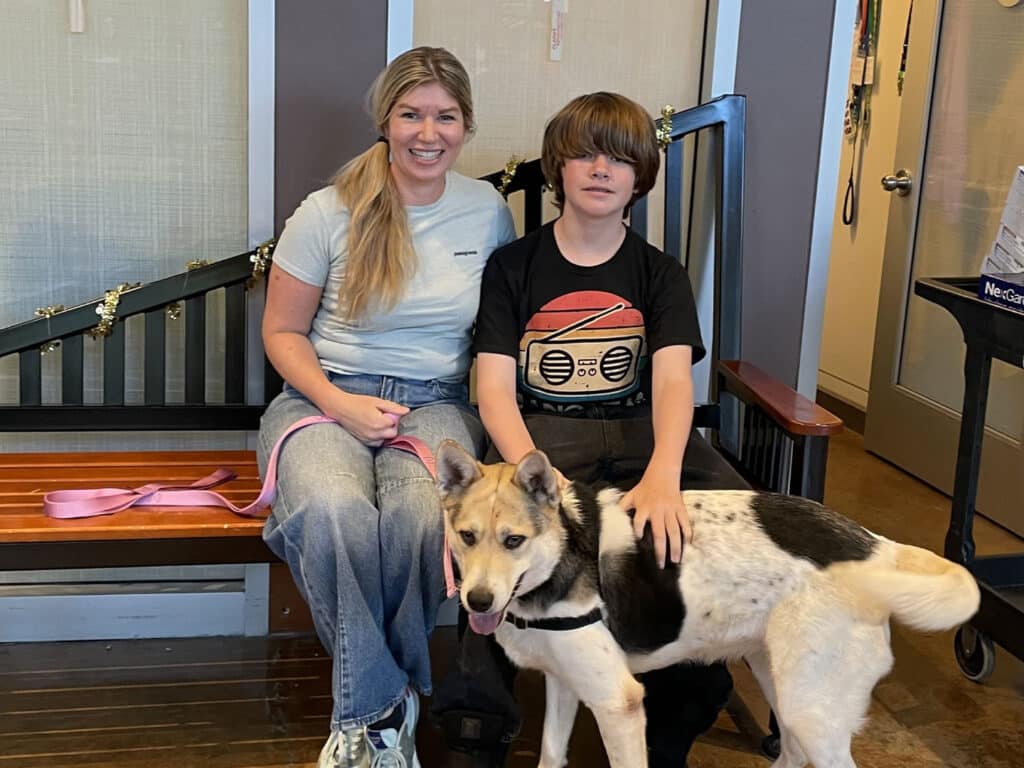 A woman and a boy are sitting on a bench inside a facility, with a dog resting on the floor beside them. The woman is wearing a light blue shirt and jeans, while the boy is in a black t-shirt and pants. They appear to be in a welcoming environment with decorative elements in the background.