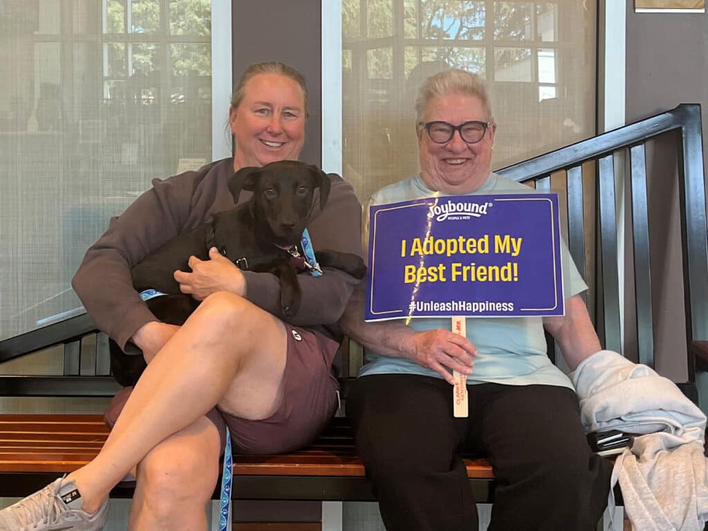 Two women sitting on a bench in a welcoming environment, holding a small black dog. One woman has a sign that reads "I Adopted My Best Friend!" The setting suggests a dog adoption event.