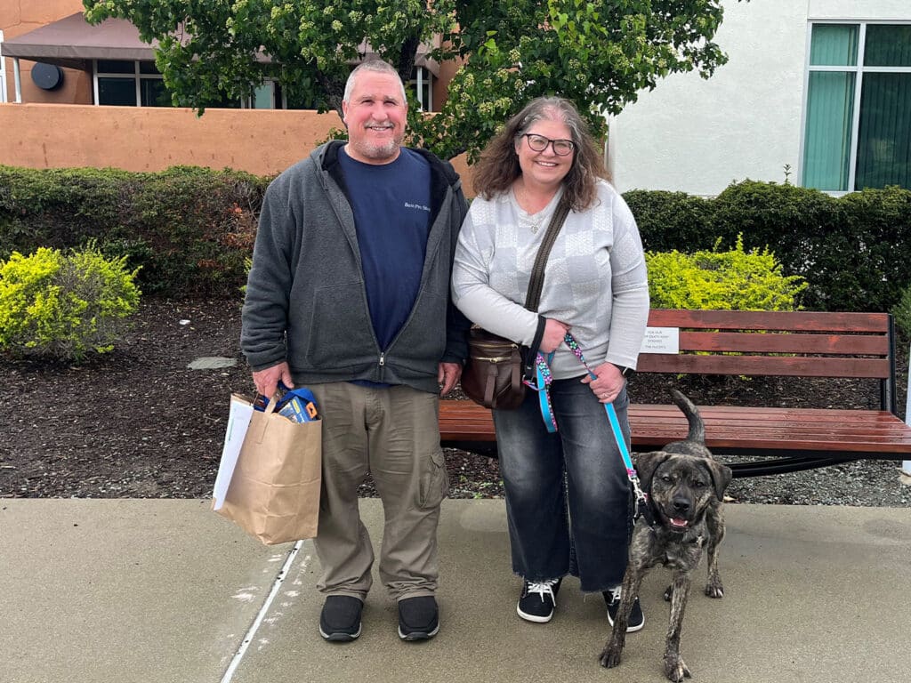 A man and a woman standing on a sidewalk beside a brown bench, smiling. The man holds a paper bag, and the woman is walking a dog on a leash. Green shrubs are in the background, and the weather is overcast.
