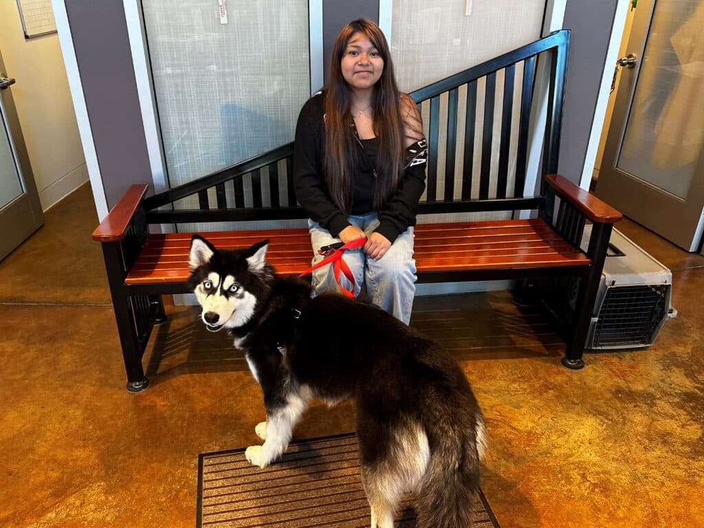 A woman with long dark hair sits on a wooden bench in an indoor space, holding a leash attached to a black and white husky dog. The background features glass doors and a patterned wall, and the floor is polished concrete.