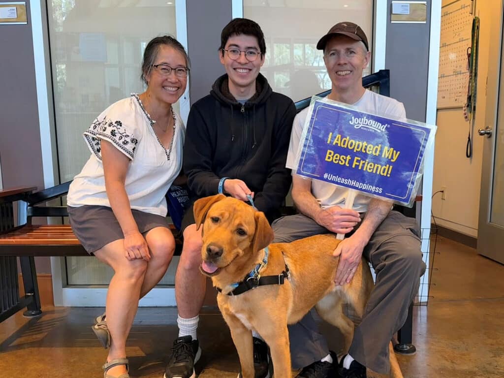 A family smiling at an adoption center, posed with a yellow Labrador. The woman on the left is wearing a white shirt and shorts, the boy in the middle is in a black hoodie, and the man on the right holds a sign that says "I Adopted My Best Friend!"
