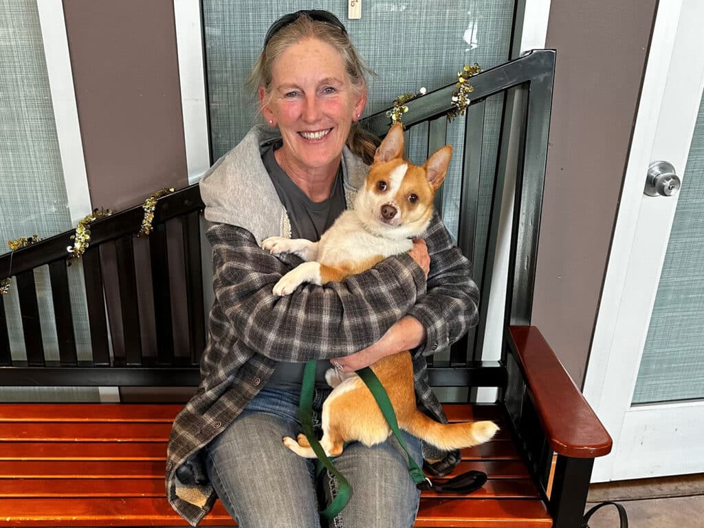 A woman sitting on a wooden bench smiles while holding a small tan and white dog in her arms. She wears a gray hoodie over a plaid shirt. In the background, there are decorations on a railing.