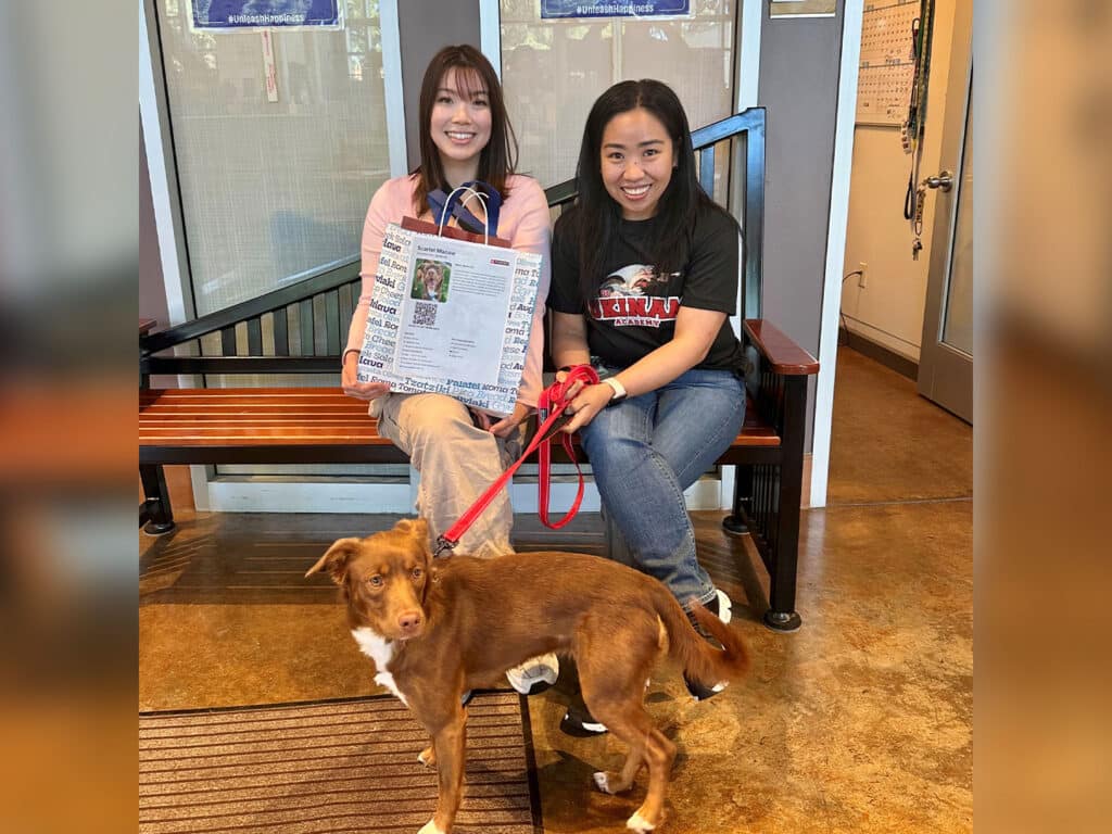 Two women sitting on a bench in a shelter, smiling and holding an adoption certificate. A brown dog is standing in front of them on a red leash, looking at the camera. The background includes shelter signage and a door.