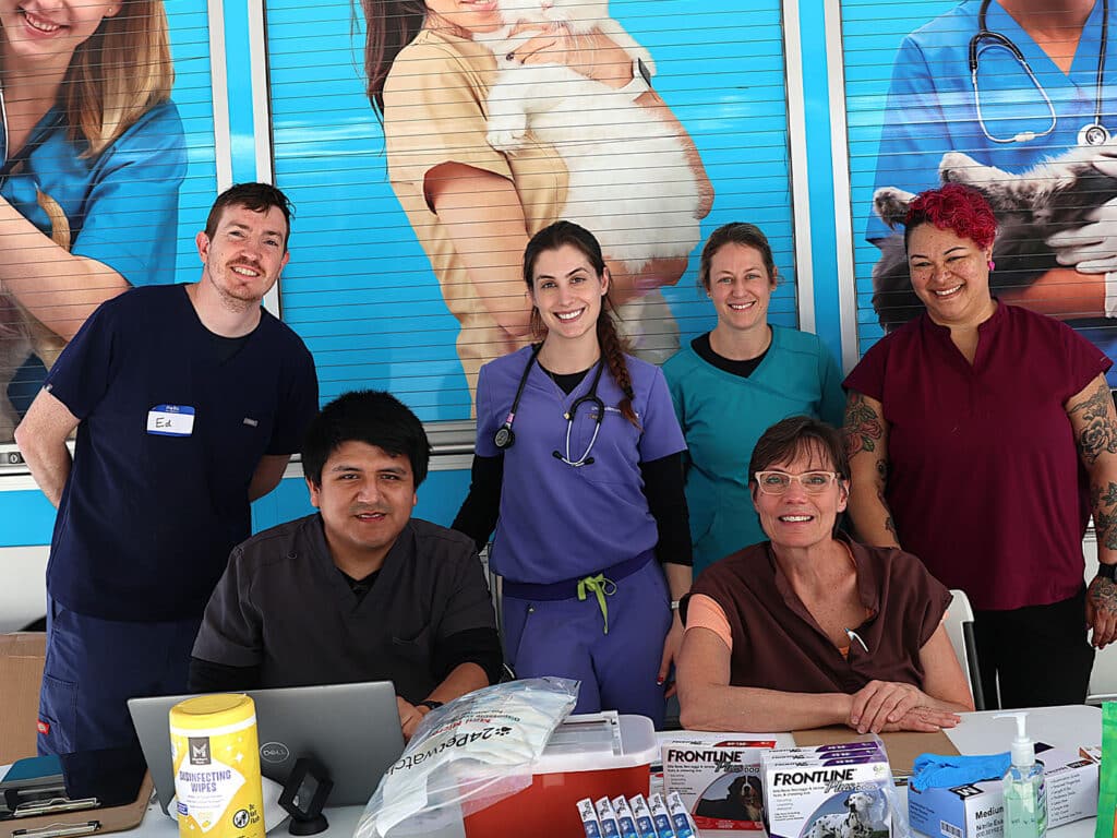 A group of six individuals posed at a community care event, with four women and two men. They are standing or sitting behind a table with pet care supplies and a laptop, smiling in front of a colorful background featuring animal care images.