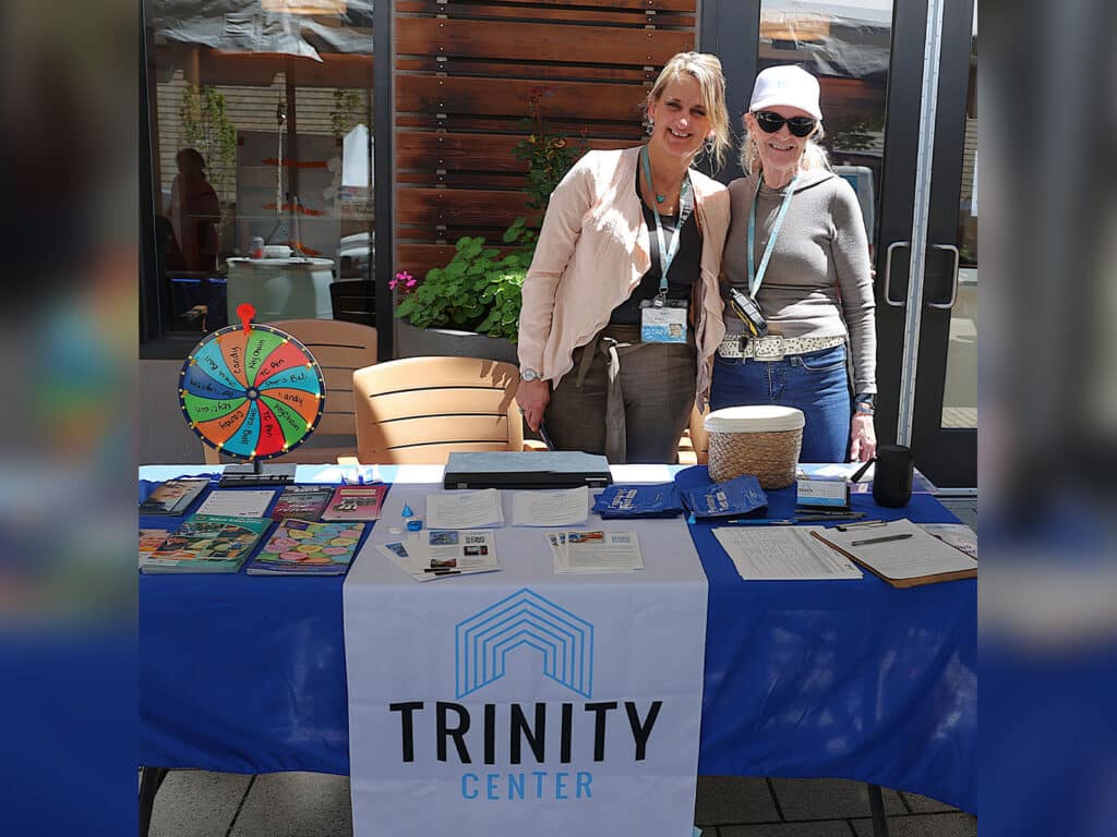 Two women standing behind a table with a blue tablecloth featuring the logo of the Trinity Center. The table displays brochures, papers, and a spinning wheel. They smile towards the camera, with a sunny outdoor setting visible in the background.