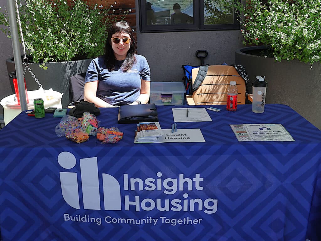 A woman wearing sunglasses sits at a table with an "Insight Housing" blue tablecloth. The table contains pamphlets, a notebook, and a variety of snacks. In the background, there are potted plants and a window.