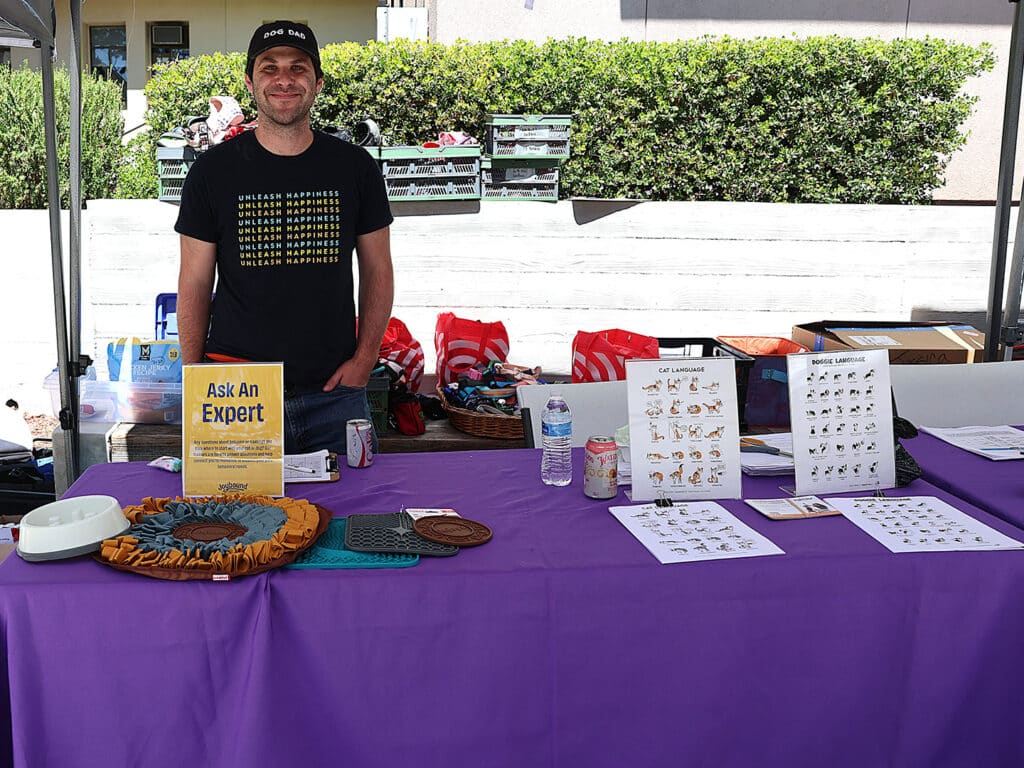 A man standing behind a purple table at an outdoor event. He is wearing a black shirt with yellow graphics. The table features informational materials, water bottles, and a sign that says "Ask An Expert." In the background, there are crates and plant greenery.