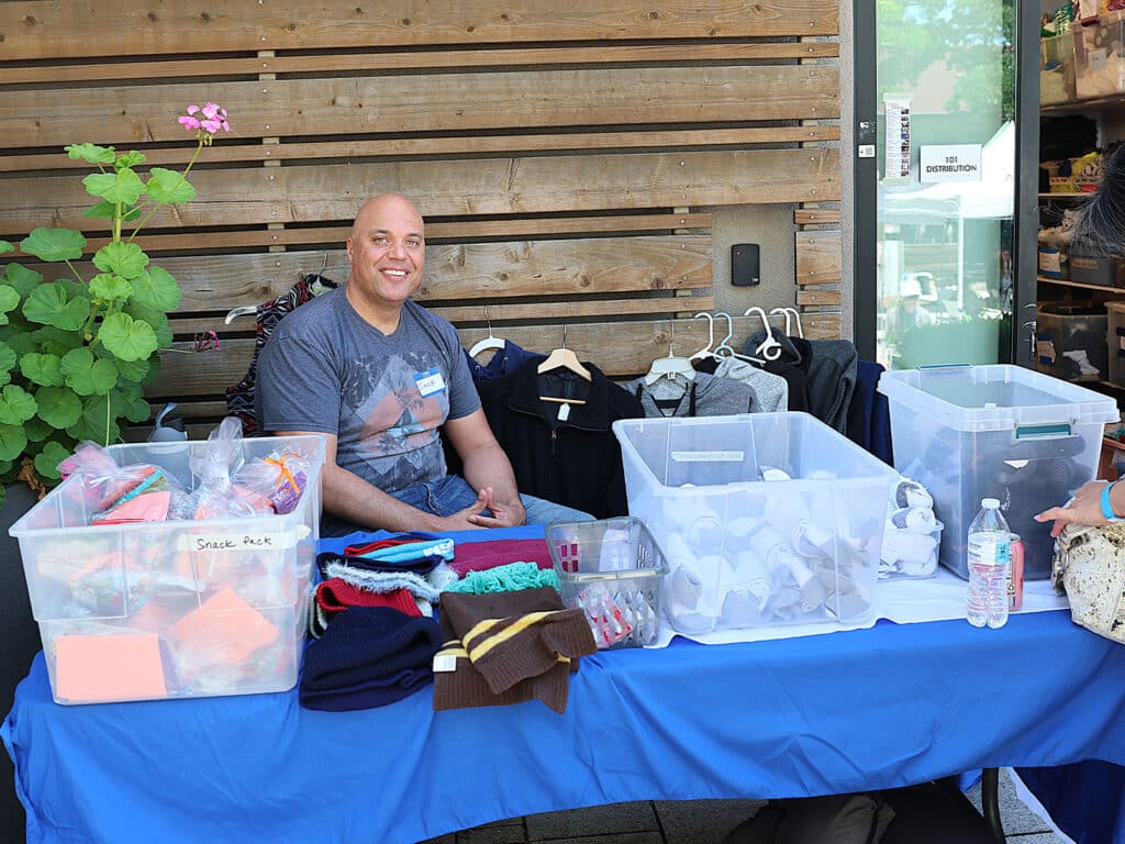 A man sitting behind a table covered with a blue tablecloth. The table displays clear storage bins filled with various items, including clothing and crafts. A green plant is visible in the background, and a building entrance can be seen on the right.