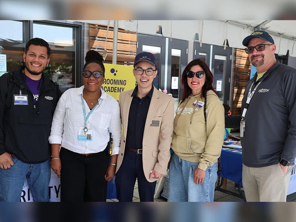 Five individuals pose together at a community event. They are standing in front of a table with informational materials, wearing name tags, and smiling. The background includes large windows and a sign for a grooming academy.
