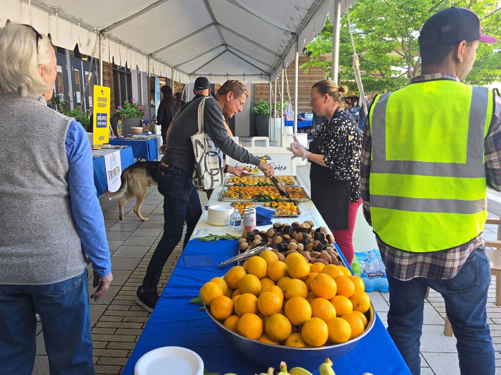 A community event under a tent featuring various food items on a long table. Individuals are serving and selecting food, including oranges in a bowl. A person in a green safety vest interacts with others. A dog is visible nearby.