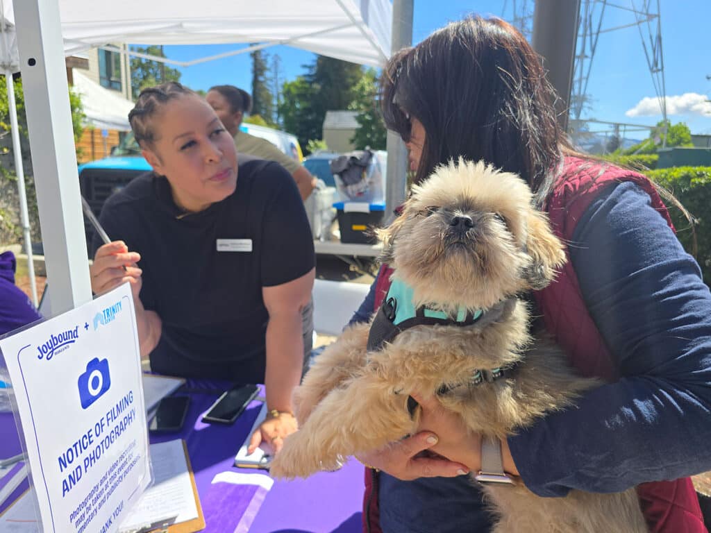 A woman holding a small dog stands next to a table with a sign that reads 'Notice of Filming and Photography.' Another woman at the table is looking at her, engaged in conversation. The setting features a clear blue sky and white canopy.