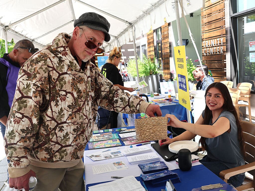 An older man in a camouflage jacket interacts with a young woman at a table during an outdoor event. The woman, wearing a dark shirt, holds a woven basket while documents and brochures are spread out on the table. People are seen in the background.