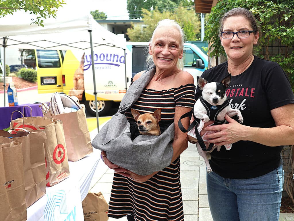 Two women smiling while holding small dogs. One woman has a Chihuahua in a gray bag, and the other holds a small dog in a harness. They stand in front of a table with bags and a vendor tent in a community outdoor setting.