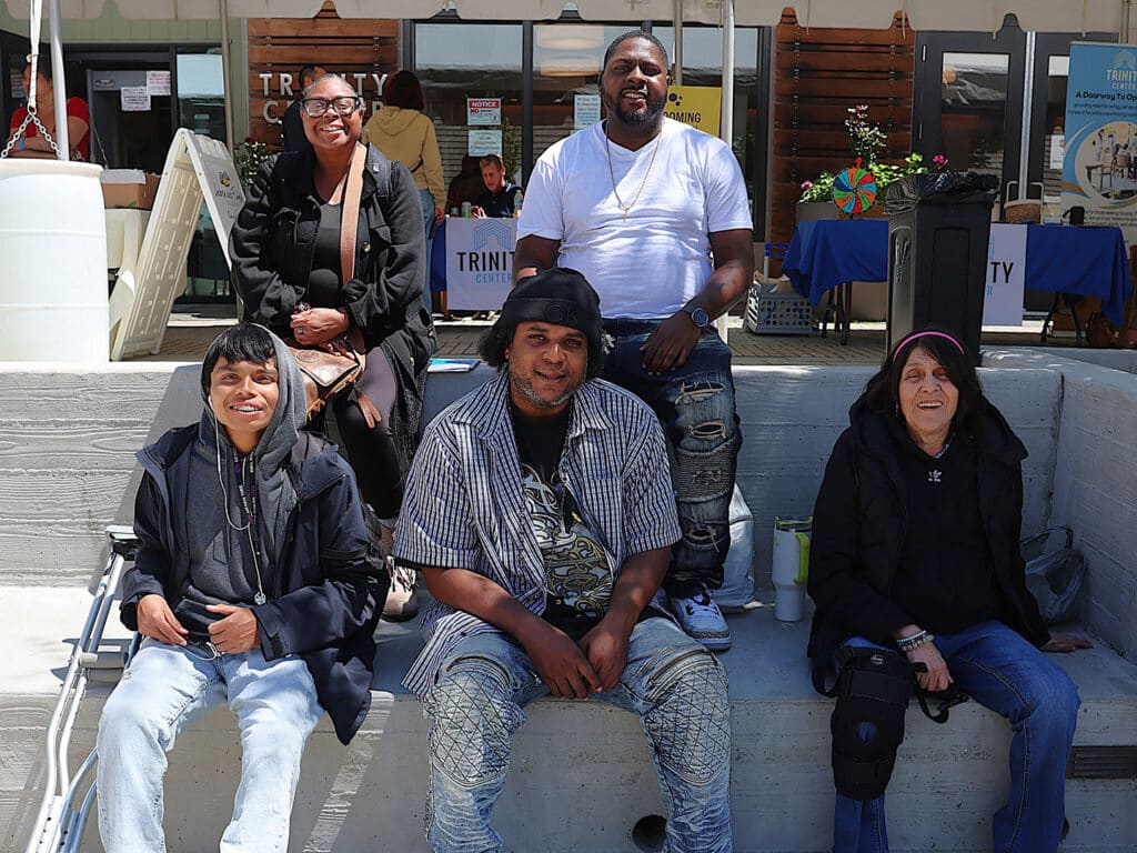 Five individuals sitting on a concrete platform outside a community center. Two women and three men are present, wearing casual clothing. A service area is visible in the background with tables and banners related to community services.