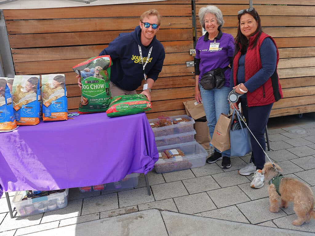 Three individuals are standing at a table covered with a purple cloth at a community event. The table displays bags of pet food and containers of snacks. A small dog is on a leash next to a woman on the right.