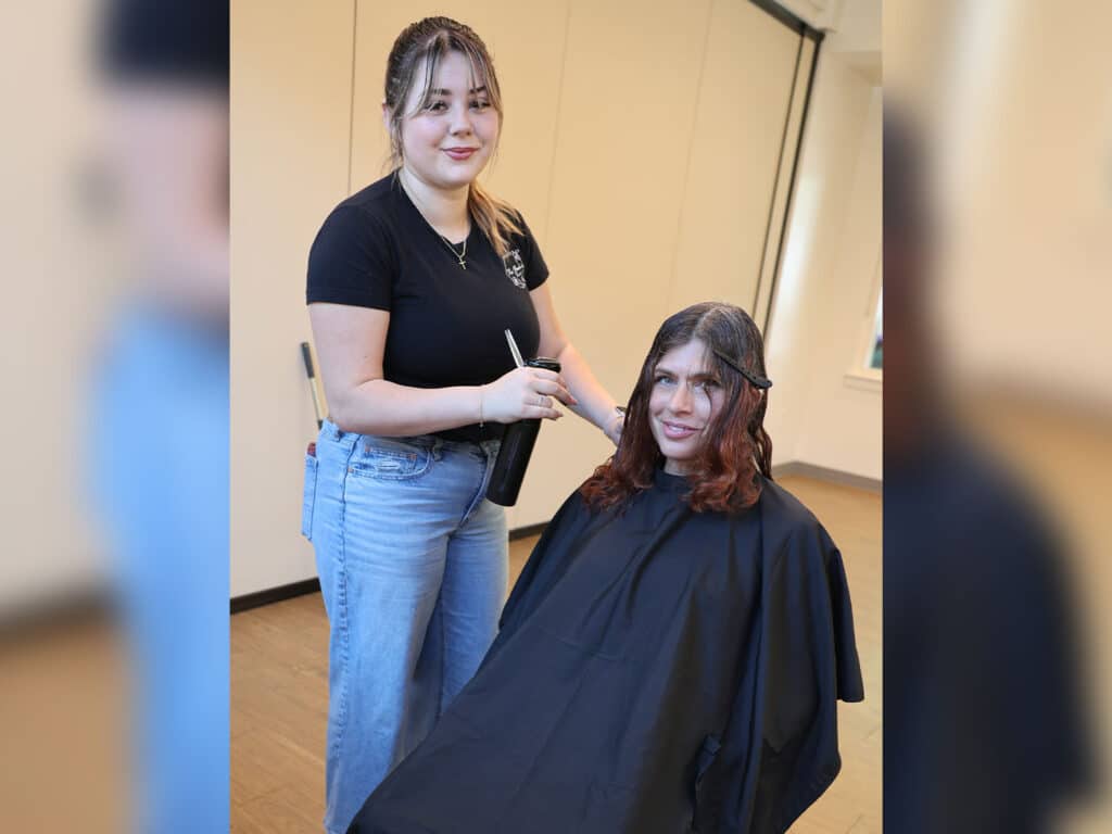 A young stylist, wearing a black shirt and blue jeans, stands beside a seated woman with long hair. The woman, covered with a black hair salon cape, is smiling as the stylist prepares to cut her hair in a bright, well-lit room.