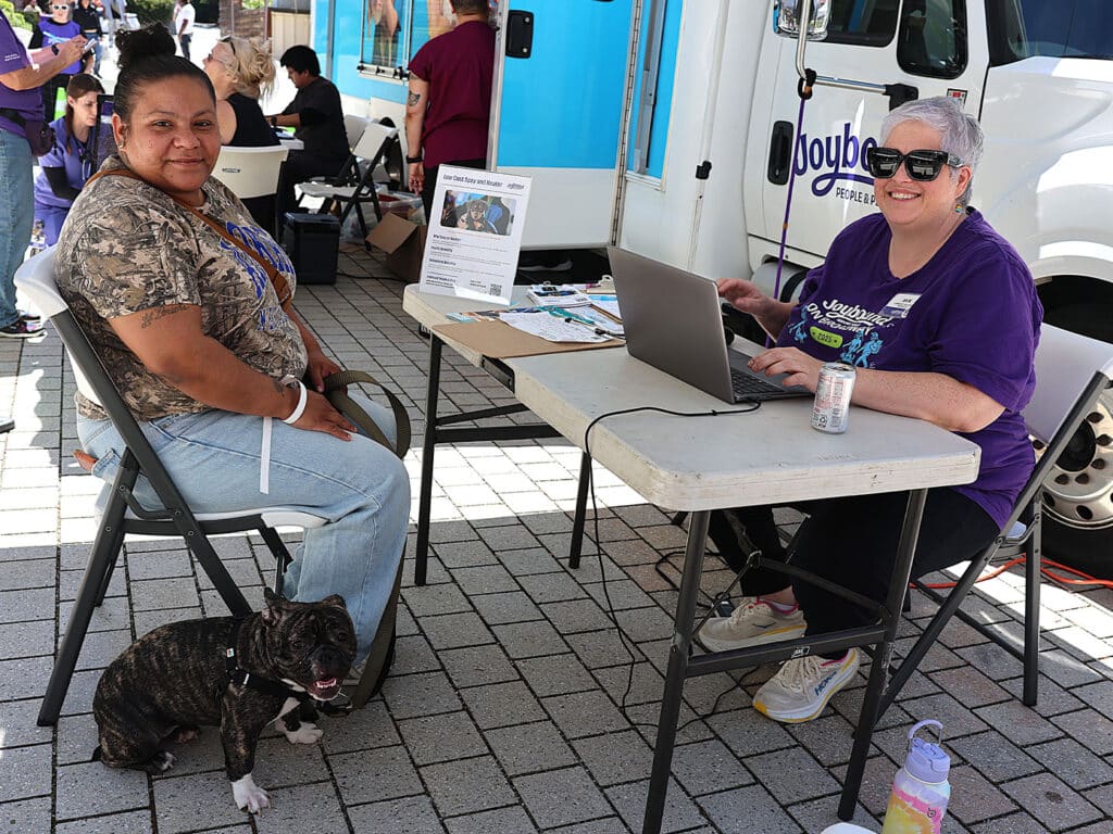 A woman in a camouflage shirt sits at a table with a small dog beside her, while another woman in a purple shirt works on a laptop. They are outdoors, with a mobile unit in the background and various items spread across the table.
