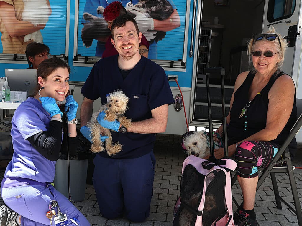 A veterinarian in scrubs holds a small dog wearing a blue outfit, while a veterinary technician kneels beside them, both smiling. An older woman sits nearby with a small dog in a pink bag. A mobile clinic is visible in the background.