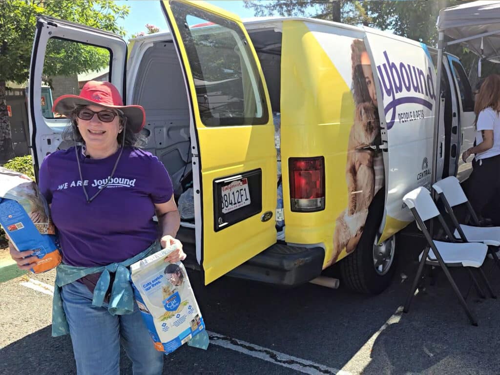 A woman wearing a purple t-shirt with the slogan "WE ARE youSound" stands outside a van with a yellow and white design. She holds bags of pet food while the van's door is open, revealing its interior. The scene is set outdoors during a sunny day.