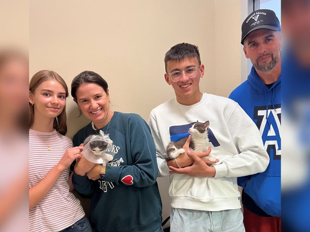 A young girl and a woman hold a cat in a clear container, while a teenage boy cradles another cat. A man stands nearby, smiling. They are inside a well-lit room, suggesting a joyful moment related to pet adoption.