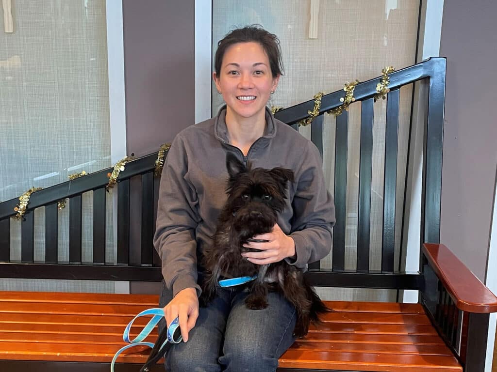 A woman sitting on a wooden bench, holding a small black dog in her lap. She wears a gray sweatshirt and has a friendly smile. The background is minimal, featuring a wall with decorations, including small golden elements.