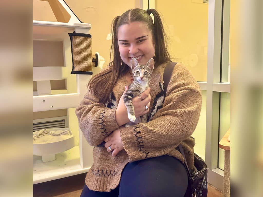 A young woman with long hair styled in two pigtails sits inside an animal shelter, holding a small gray and white kitten. She wears a cozy sweater and smiles at the camera, surrounded by a light-colored shelter interior with cat structures in the background.