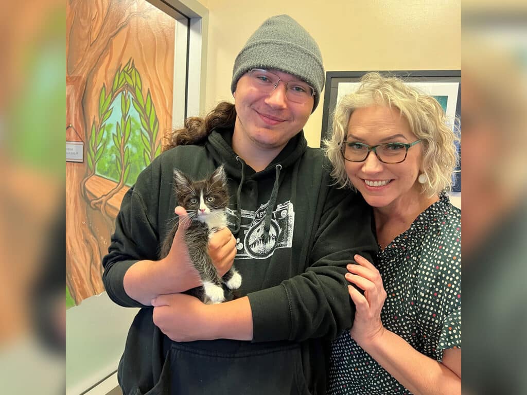 A young man in a gray beanie holds a small gray and white kitten with green eyes, while a woman with short blonde hair and glasses stands beside him. They both smile at the camera in a room featuring colorful wall art.