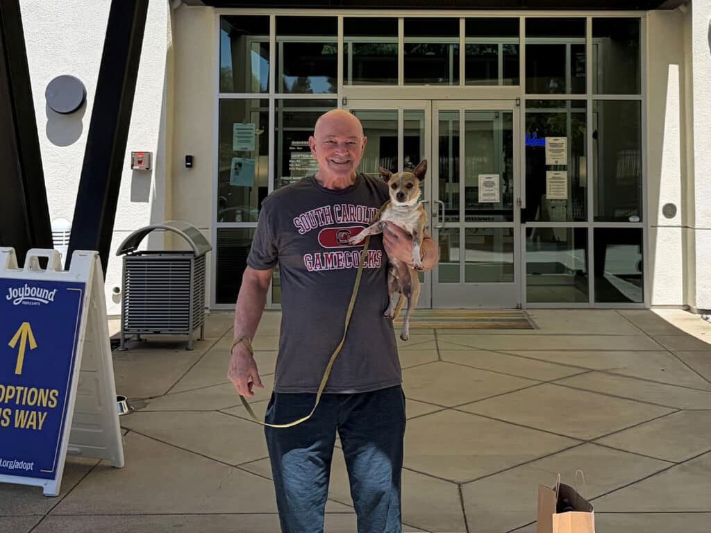 A man with a shaved head, wearing a gray t-shirt that says 'South Carolina Gamecocks,' holds a small dog under his arm outside an adoption center. The building features large glass doors and a sign pointing towards options.