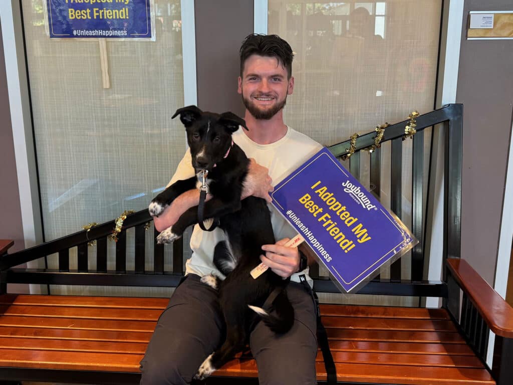 A man sitting on a bench inside an adoption center, holding a black dog in his lap. The man is smiling and wearing a white polo shirt. He is displaying a sign that reads "I Adopted My Best Friend!" in blue and yellow text.