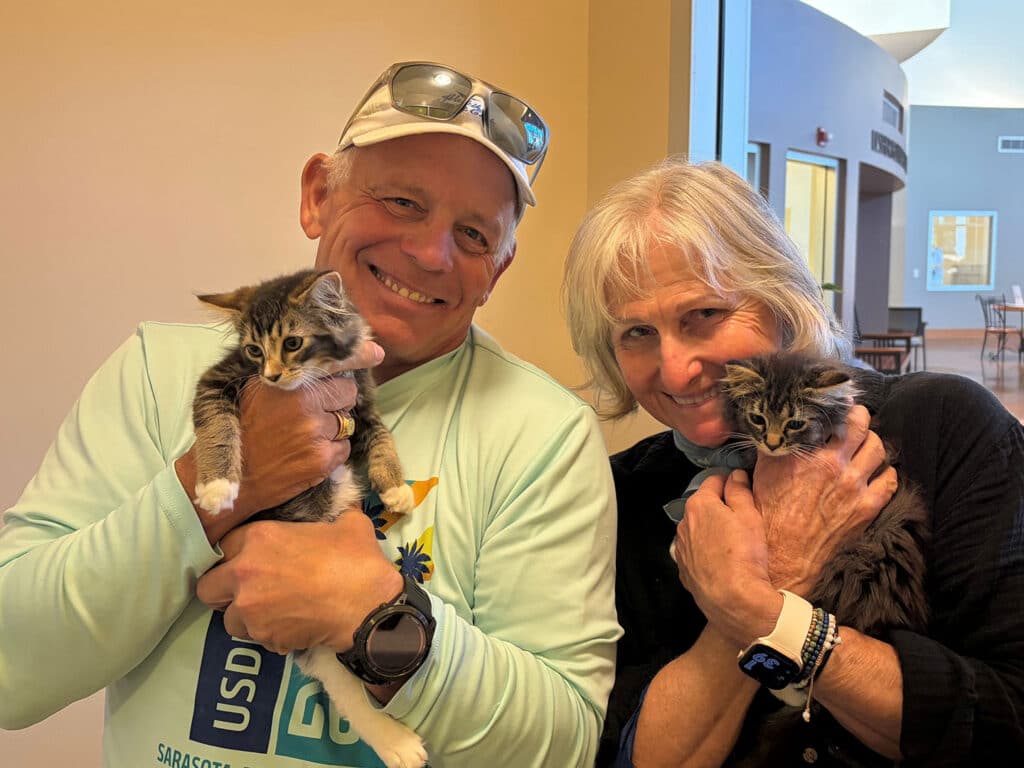 A man and a woman smile while holding two fluffy kittens in their arms. The man wears a light blue shirt and sunglasses on his head; the woman has long hair and wears a black jacket. They are inside a brightly lit room with large windows.