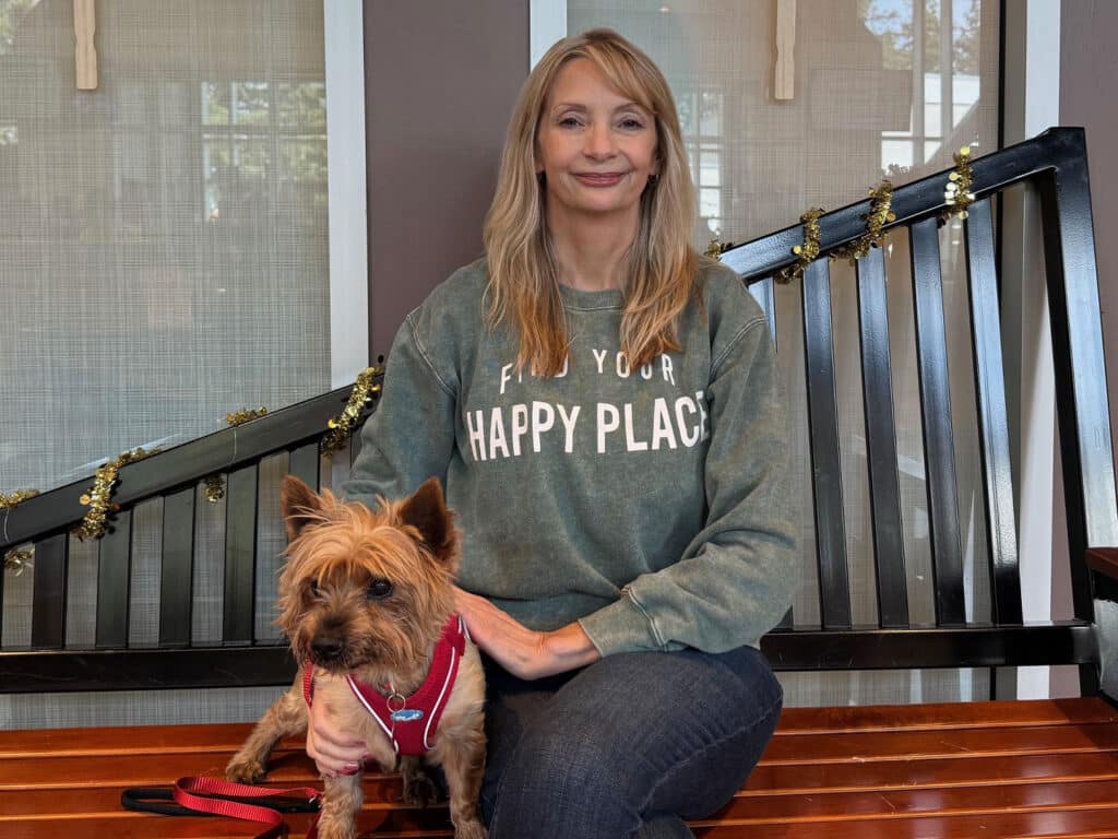 A woman seated on a bench, wearing a gray sweatshirt that reads "Find Your Happy Place," beside a small tan dog wearing a red harness. They are in a well-lit indoor space with a wooden bench and decorative accents in the background.