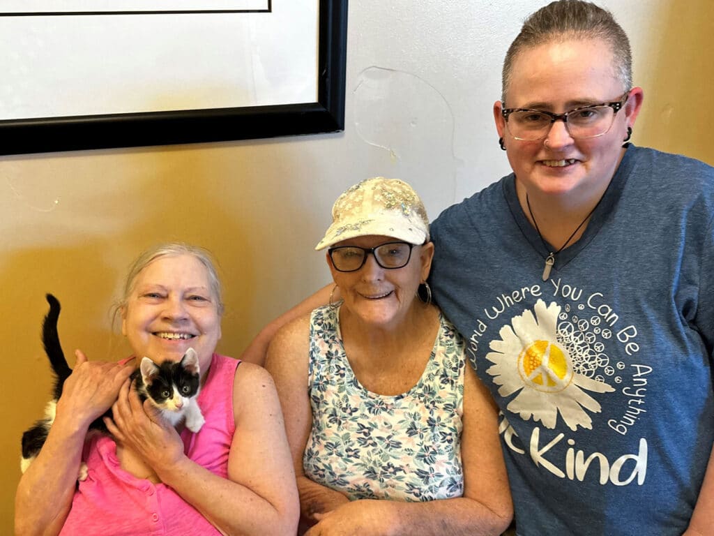 Three women are smiling together in a room. The woman on the left holds a small black and white kitten. The woman in the middle wears a white cap and a floral top, while the woman on the right wears glasses and a blue t-shirt with a message about kindness.