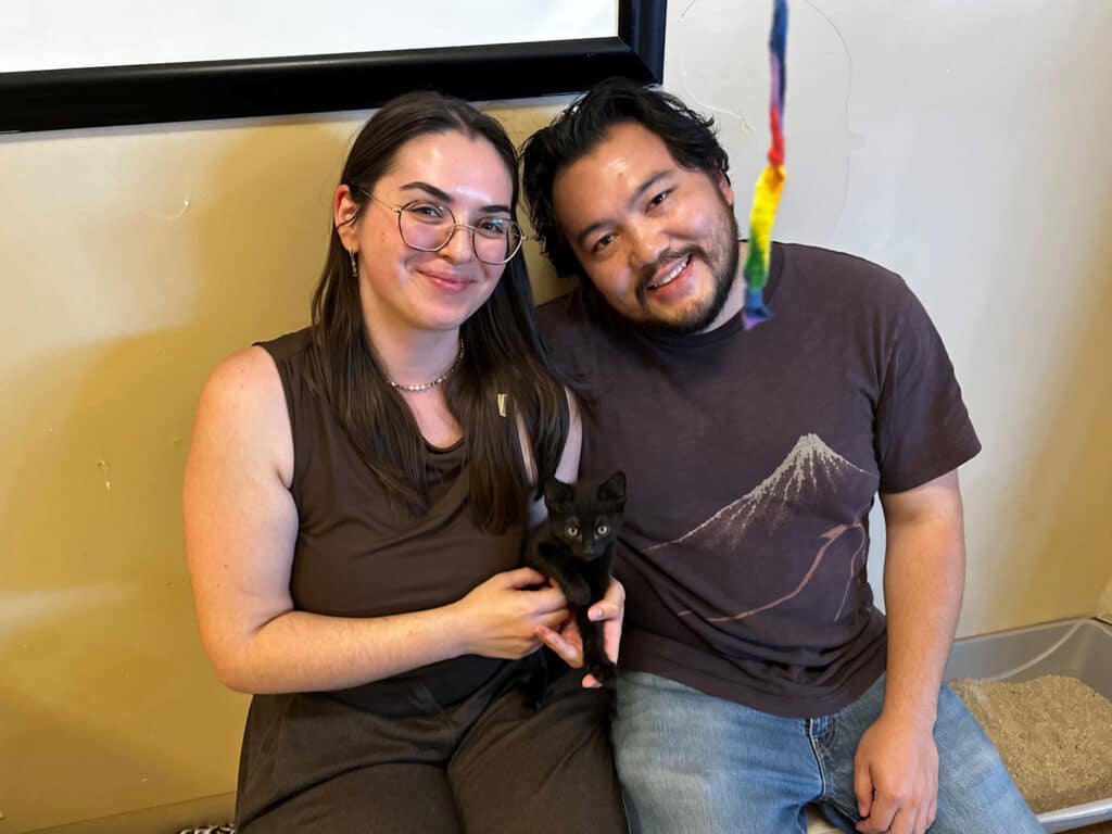 A woman and a man sitting on a beige carpet, holding a small black kitten between them. The woman is wearing a brown outfit with glasses, while the man is in a dark t-shirt and light blue jeans. They smile at the camera, with a colorful toy hanging in the background.