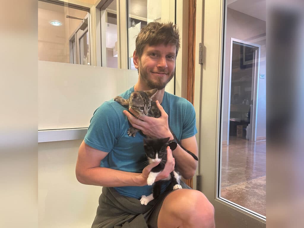 A man sitting on the floor against a glass door, holding two kittens—one gray and white on his left shoulder, and a black and white one in his right hand. He is wearing a blue t-shirt and gray shorts, smiling gently.