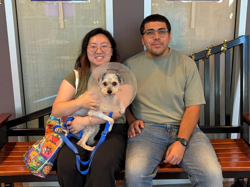 A woman and a man sit on a wooden bench, smiling with a small dog wearing an Elizabethan collar. The woman holds the dog, which is secured with a blue leash, while both wear casual clothing. The background is lightly lit, creating a welcoming setting.