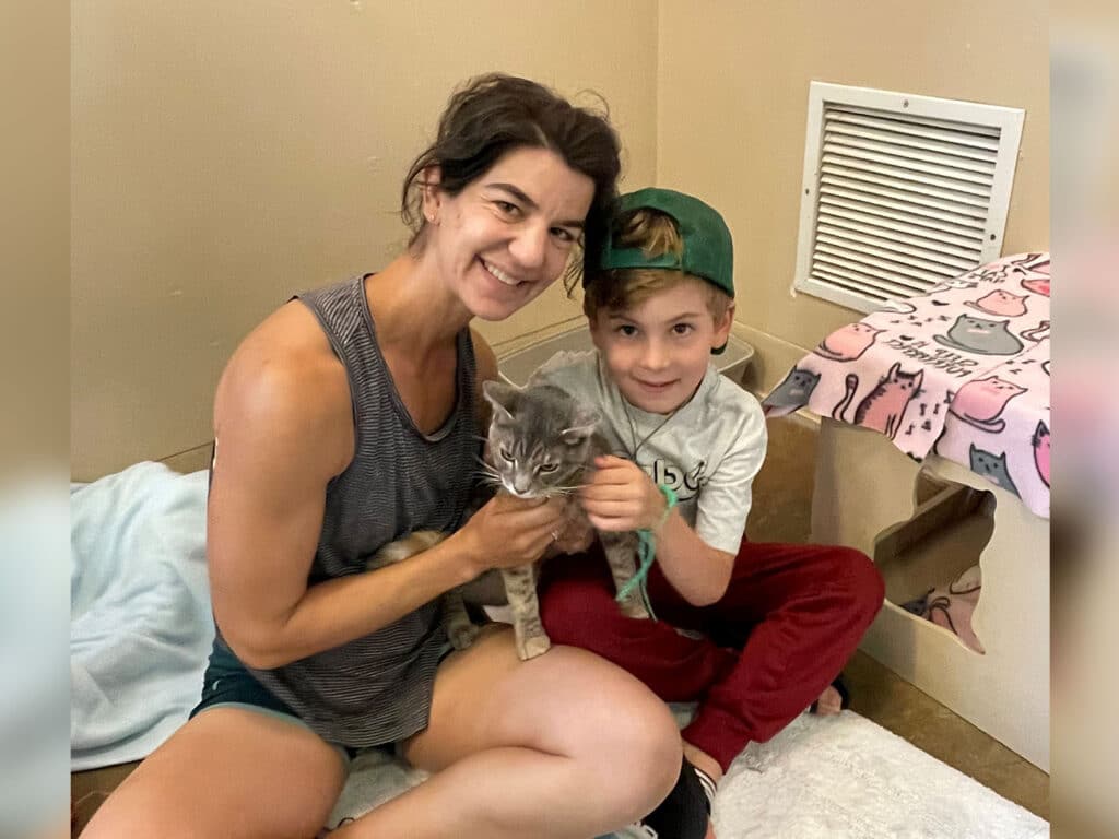 A woman and a young boy sit on the floor of an animal shelter room. The woman holds a gray cat while the boy smiles beside her. The room features a cozy setting with a patterned blanket and an air vent on the wall.