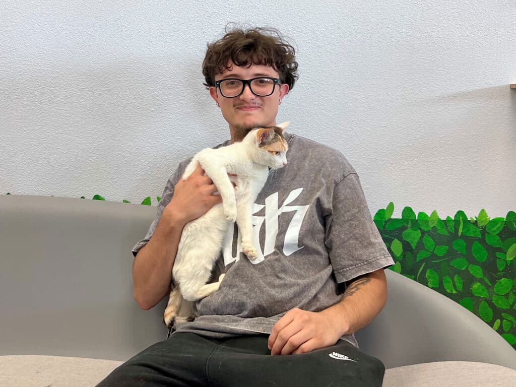 A young man with curly hair and glasses sitting on a light-colored couch, holding a white cat with orange markings. The background features a green design resembling leaves.
