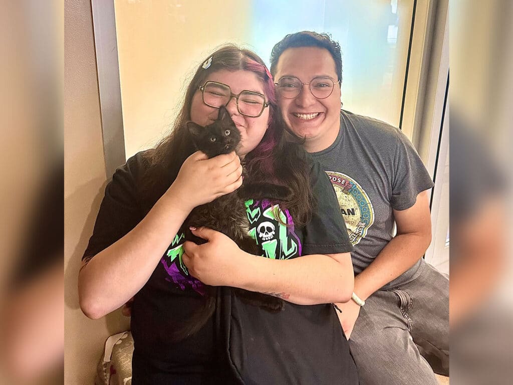 A woman with glasses holds a black cat close to her face while smiling, sitting next to a man who also has glasses and is smiling. They are in a light-colored room with neutral walls, appearing cheerful together.