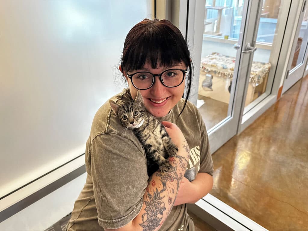 A woman with dark hair and glasses is smiling while holding a gray tabby cat against her chest in an indoor space. The background features glass doors and a clean, modern environment, suggesting a pet adoption setting.