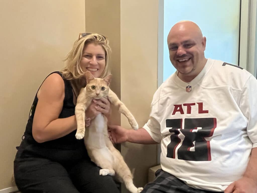 A woman with blonde hair holds a light-colored cat while sitting next to a man with a shaved head wearing an Atlanta Falcons jersey. They are smiling and seated in a brightly lit room with beige walls.