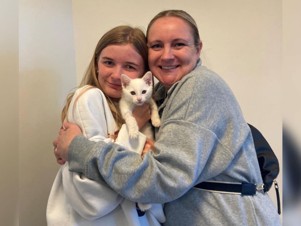 Two women smiling while holding a white kitten, standing closely together in a well-lit indoor space. One woman has long brown hair and is wearing a light gray outfit, while the other woman has blonde hair and is dressed in a white sweatshirt.