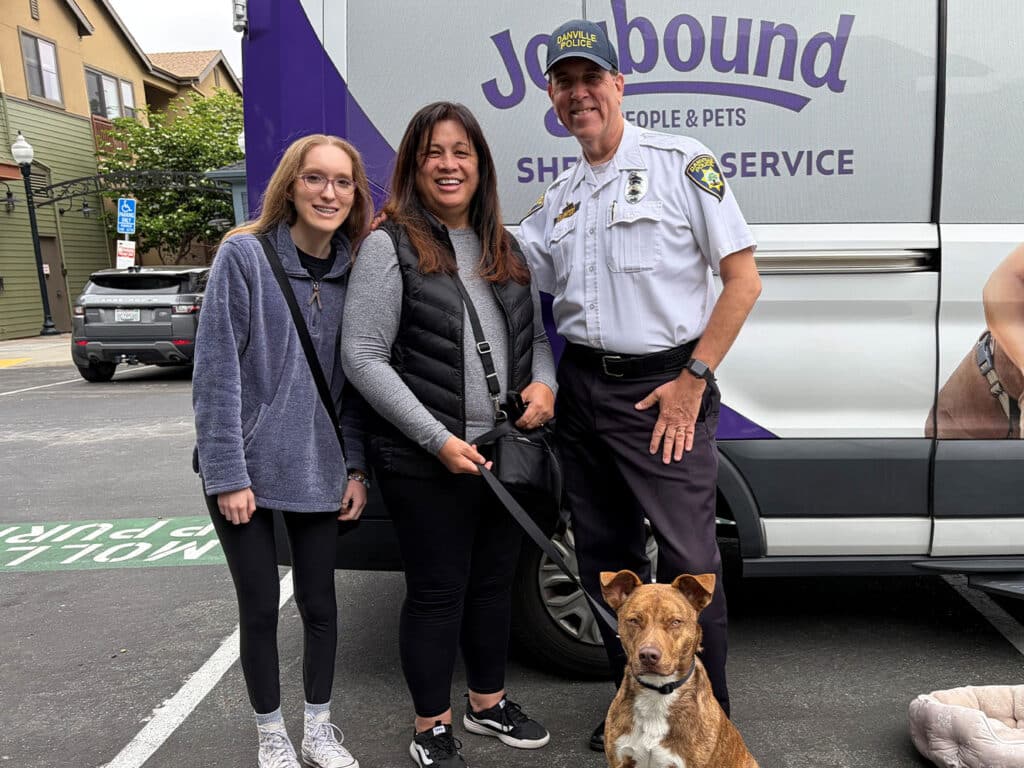 A woman with long dark hair, a girl with glasses, and a man in a police uniform pose together outside a van marked "Jobbound: People & Pets". The woman holds a dog, a light brown and white mixed breed, standing next to her.