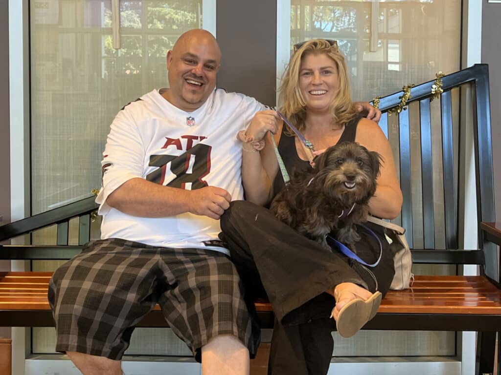 A man and a woman sitting on a bench in a lobby, smiling and holding a small black dog. The man is wearing a white shirt with black and red accents and black plaid shorts, while the woman is dressed in black pants and a black top.