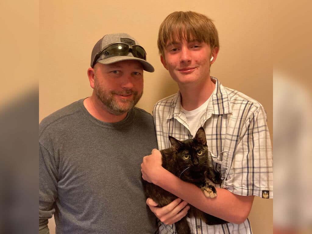 A man wearing a gray shirt and sunglasses stands beside a teenage boy in a plaid shirt. The boy holds a black and brown tortoiseshell cat. They both smile in front of a light-colored wall, showing a moment of companionship.