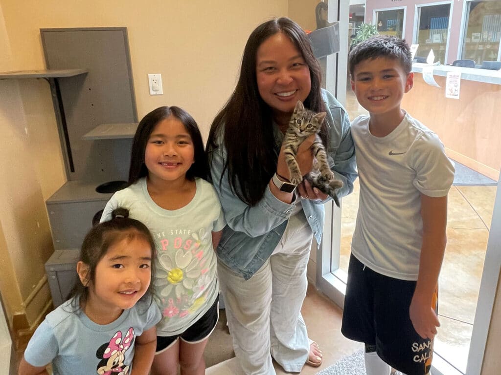 A woman holds a gray tabby kitten while standing with three children in a brightly lit room. The children, two girls and a boy, are smiling and positioned beside her, with various animal adoption displays visible in the background.