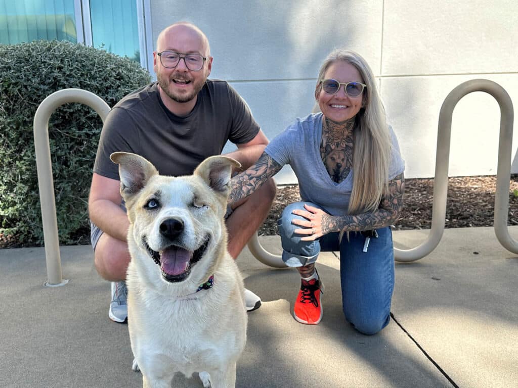 A smiling couple poses with a happy dog in an outdoor setting. The man is sitting on the ground on the left, wearing a gray shirt. The woman is kneeling on the right, wearing glasses and a gray shirt, with tattoos visible on her arms. The dog is a cream-colored mixed breed with one eye.