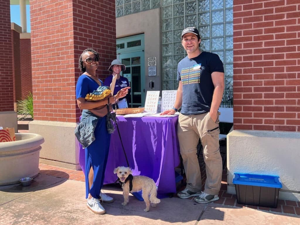 A woman in blue scrubs holding a basket stands next to a man in a black shirt and khaki pants. They are beside a table covered with a purple cloth, outdoors, with a small dog at their feet.