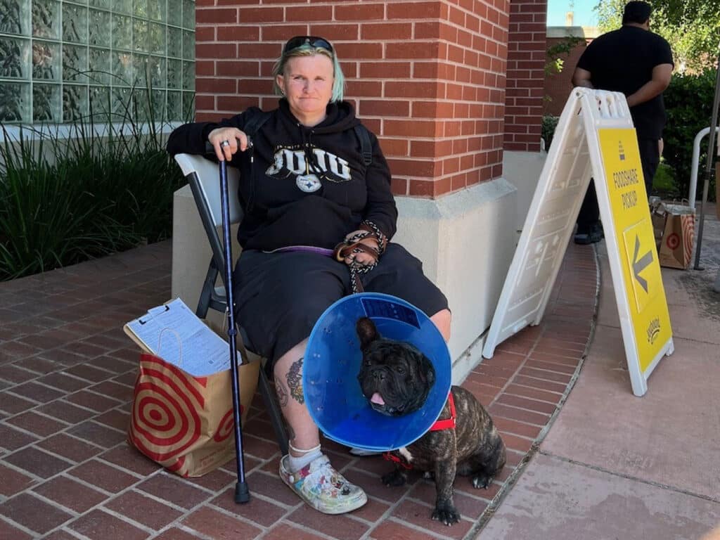 A woman sits on a chair outside a building, wearing a black hoodie and holding a cane. Beside her is a French Bulldog wearing a blue cone collar. A bag and paperwork are placed on the ground next to her, with a sign for a food service visible nearby.