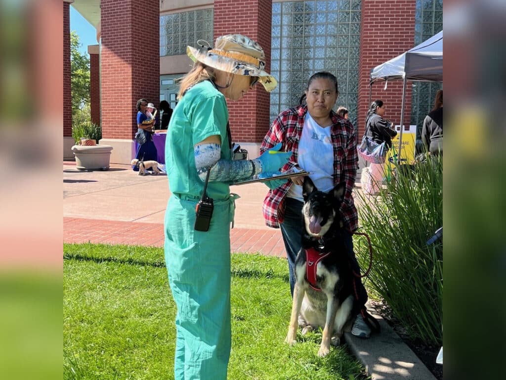 A veterinarian in green scrubs and a sunhat holds a clipboard while interacting with a woman in a red and black plaid shirt, who is standing beside a dog. The setting is outdoors, near a brick building with people in the background.