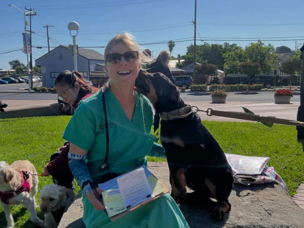A veterinarian in green scrubs sits on a stone, smiling while a dog licks her face. In the background, a person with a small dog is seated on the grass, and there are trees and buildings in view under a blue sky.